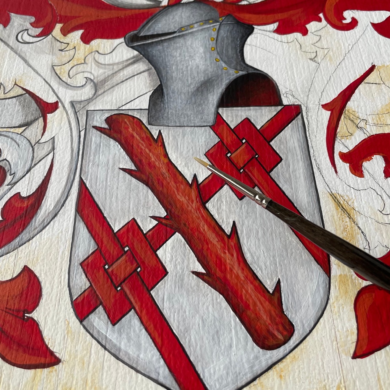 Detail of a hand-painted red heraldic shield in progress, showing an interlaced cross design and the painter's brush at work