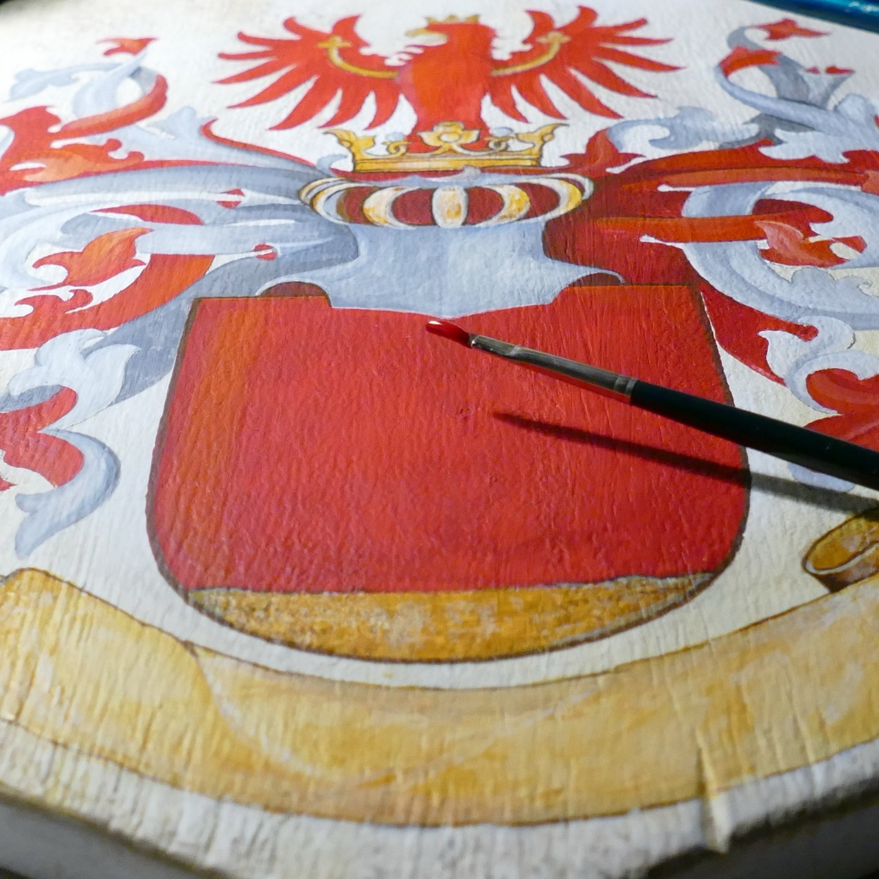 Detail of a hand-painted heraldic shield in progress, showing a red field beneath a crowned helm